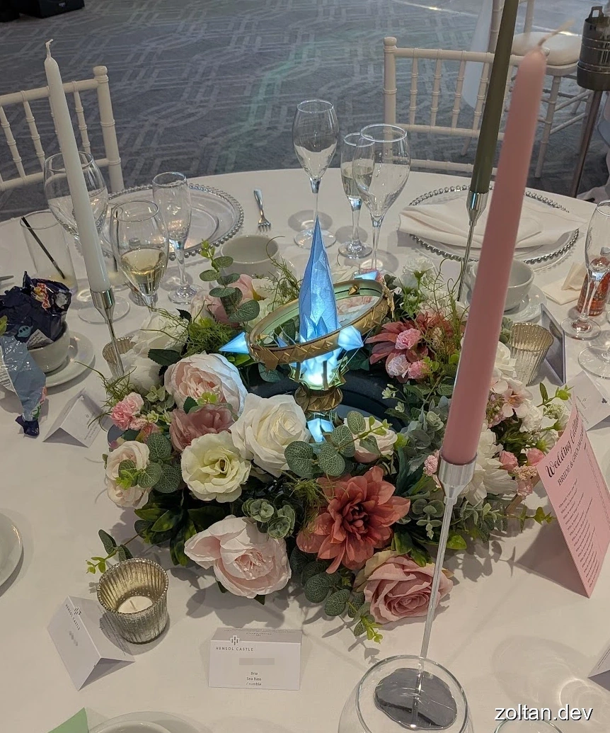 A beautifully arranged table centerpiece featuring a glowing blue Aetheryte Crystal model surrounded by flowers and candles in white and pink, set against a backdrop of elegant glassware and place settings.