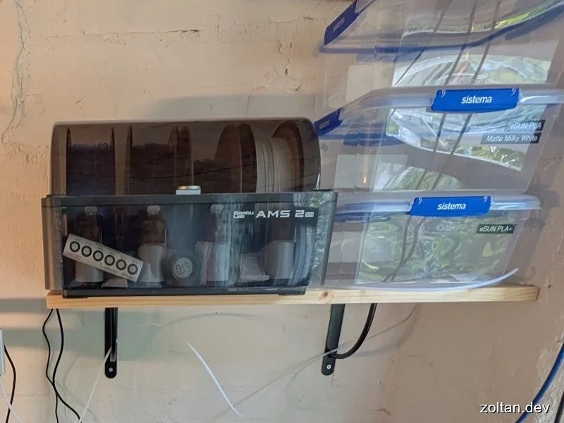 Wall-mounted shelf holding a transparent AMS2 printer enclosure and plastic storage containers labeled "Sistema" containing green filament spools.