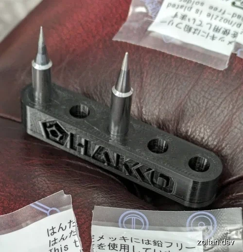 HAKKO branded soldering iron tips in a 3D-printed holder with packaging and labels visible on a textured surface.