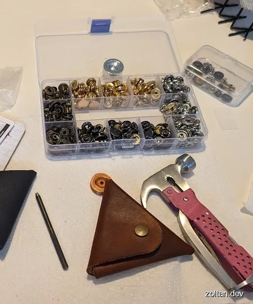 A clear box filled with various metal snap fasteners, a hammer and a laser cut leather triangle coin purse.