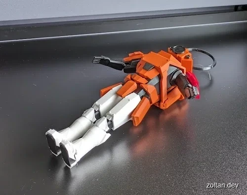 An orange and white 3D-printed robot model lies on a dark surface, showcasing intricate mechanical details and a keychain loop attached to its head.