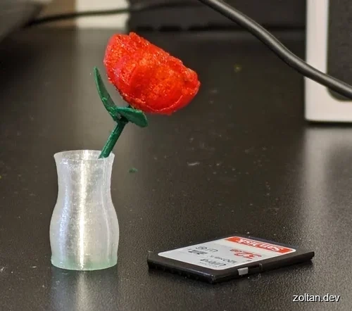 A miniature 3D-printed red carnation in a clear 3D printed vase sits next to an SD card on a dark surface.