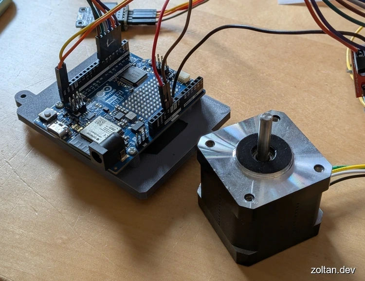 Arduino board connected to a stepper motor via wires and 3D-printed brackets on a wooden surface.