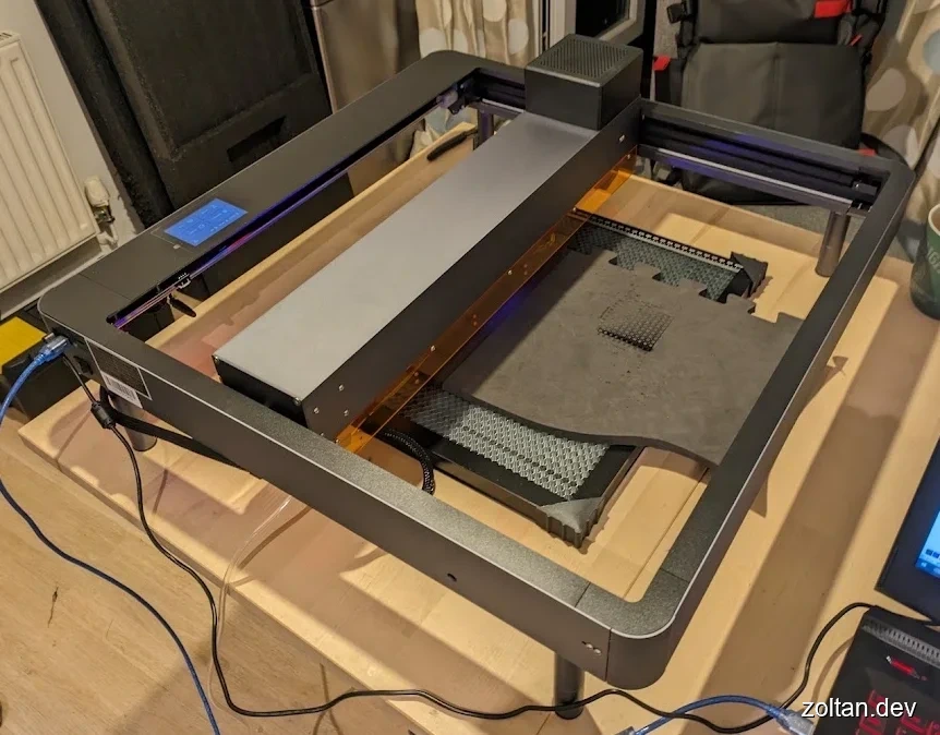 A 3D printer setup on a wooden table with visible components like the build platform, extruder, and control panel, showcasing a workspace for 3D printing projects.
