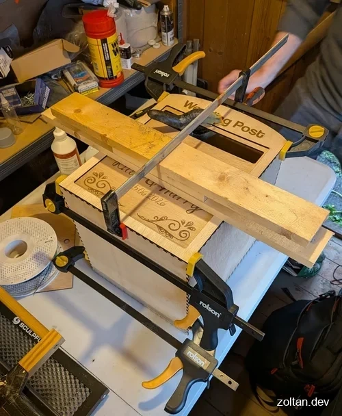 Workshop scene featuring a wooden mailbox being sanded and clamped in place, surrounded by various tools and supplies on a workbench.