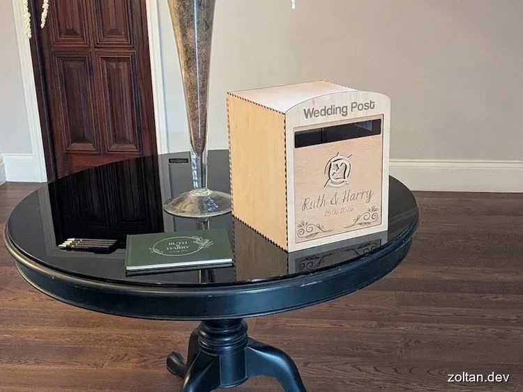 A wooden wedding post box sits atop a round black table alongside a green guest book and pens, with a decorative vase in the background.